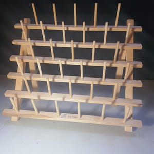 May include: A wooden thread rack with multiple rows of pegs for holding spools of thread. The rack is made of light-colored wood and has a rectangular shape. The brand name "June Tailor" is visible on the bottom shelf.