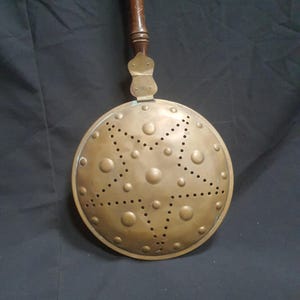 May include: Antique brass bed warmer with a wooden handle. The round warming pan features a pattern of raised circles and small holes. The handle is dark brown and attached to the pan with a brass fitting.
