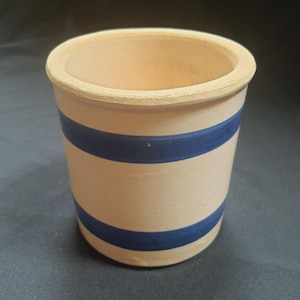 May include: A cylindrical, off-white ceramic pot with two horizontal blue stripes. The rim and base have a slightly rough texture. The pot is likely for kitchen or home decor use.