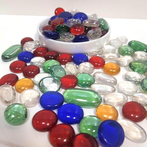 May include: A collection of smooth, rounded glass pebbles in various colors including red, blue, green, yellow, and clear. Many of the pebbles are scattered across the surface, while others are contained within a small, round white bowl.