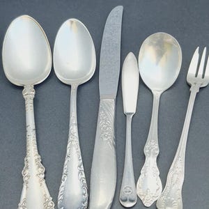 May include: A set of six silver-plated utensils, including a butter knife, a table knife, a soup spoon, a teaspoon, a fork, and a dessert spoon. The utensils have ornate handles with floral designs.