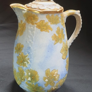 May include: A vintage ceramic pitcher with a lid, featuring a light blue background and yellow floral patterns. The pitcher has a curved handle and a gold-trimmed rim. The lid has a loop handle. A decorative piece for serving beverages.