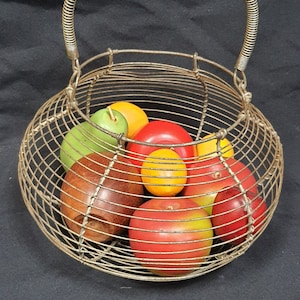 May include: A wire basket with a handle, filled with faux fruit. The fruit includes red, yellow, green, and brown apples.