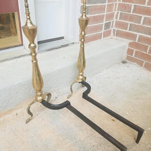 May include: A pair of antique brass and black fireplace andirons. The andirons have a tall, ornate brass design with a pointed top and a curved base. The base is black and has a long, straight bar that extends out to hold the logs.