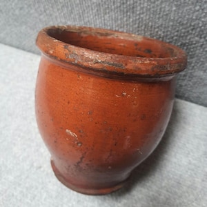 May include: A small, antique terracotta-colored ceramic pot with a rounded body and a slightly flared rim. The pot has a weathered appearance with visible cracks and imperfections, suggesting age and use. The rim shows signs of wear, adding to its vintage charm.