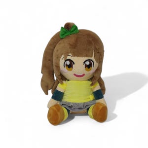 Puede incluir: Una mu&ntilde;eca de peluche con cabello casta&ntilde;o, un lazo verde y ojos grandes y expresivos. La mu&ntilde;eca lleva una camisa amarilla, una falda gris y zapatos marrones. La mu&ntilde;eca est&aacute; sentada.