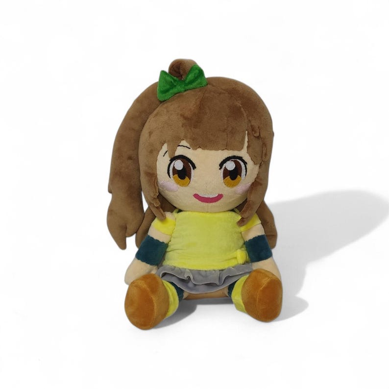 Puede incluir: Una mu&ntilde;eca de peluche con cabello casta&ntilde;o, un lazo verde y ojos grandes y expresivos. La mu&ntilde;eca lleva una camisa amarilla, una falda gris y zapatos marrones. La mu&ntilde;eca est&aacute; sentada.
