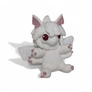 Puede incluir: Un peluche blanco con forma de criatura fant&aacute;stica con ojos grandes y redondos de color burdeos y peque&ntilde;as alas. El juguete tiene detalles en rosa en las orejas, las patas y las puntas de las alas. La criatura est&aacute; sentada con los brazos extendidos.