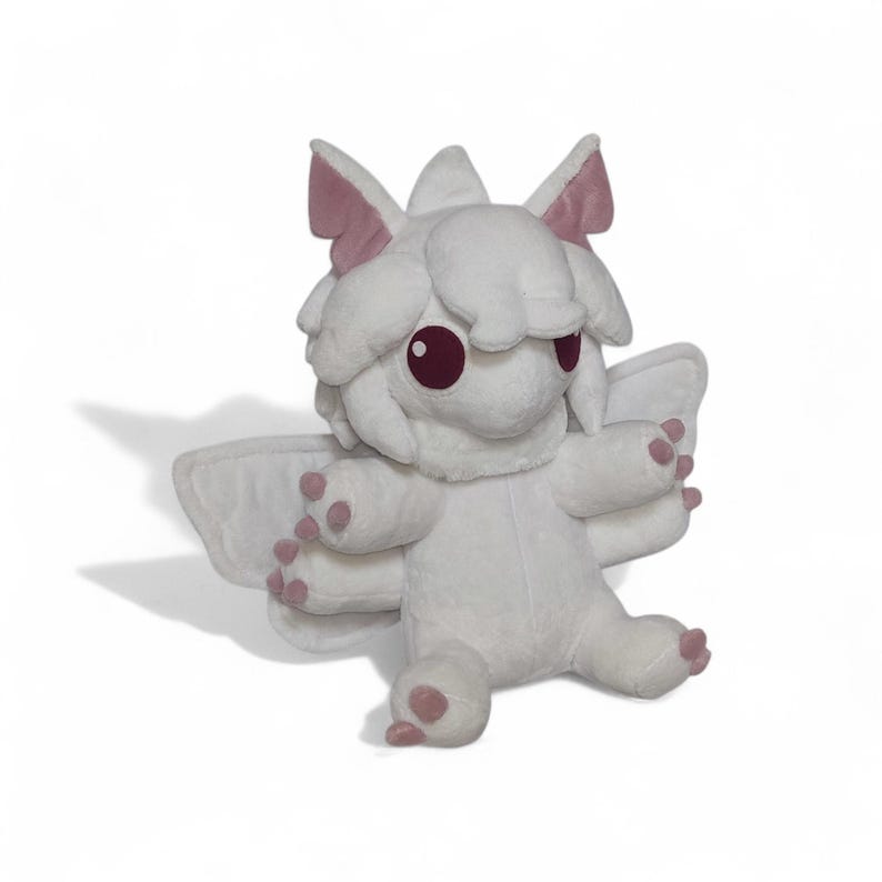 Puede incluir: Un peluche blanco con forma de criatura fant&aacute;stica con ojos grandes y redondos de color burdeos y peque&ntilde;as alas. El juguete tiene detalles en rosa en las orejas, las patas y las puntas de las alas. La criatura est&aacute; sentada con los brazos extendidos.