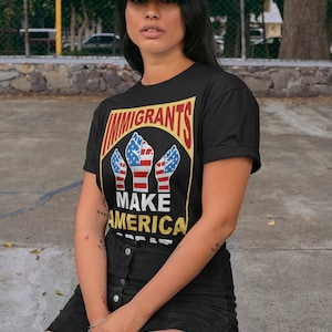 May include: A black t-shirt with a graphic of three raised fists with the American flag design. The text "Immigrants Make America Great" is printed on the shirt. The person is wearing a black baseball cap with the text "Make America Kind Again" printed on it.