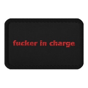 May include: Black rectangular patch with the embroidered phrase "fucker in charge" in red. The patch has a stitched border and a textured appearance, suitable for application on clothing or accessories.