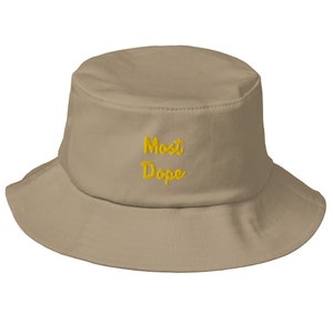 Puede incluir: Un sombrero de pescador beige con la inscripción bordada "Most Dope" en amarillo. El sombrero tiene un ala ancha y un diseño sencillo, adecuado para ropa informal. El sombrero está hecho de un material suave.