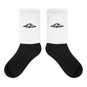May include: A pair of white crew socks with black heels and toes. The socks feature a black and white graphic of a UFO on each sock.