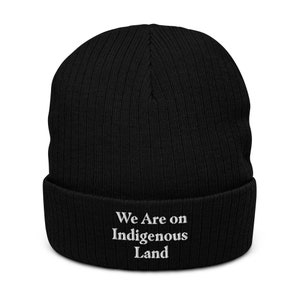May include: Black knitted beanie with a white embroidered message that reads "We Are on Indigenous Land".