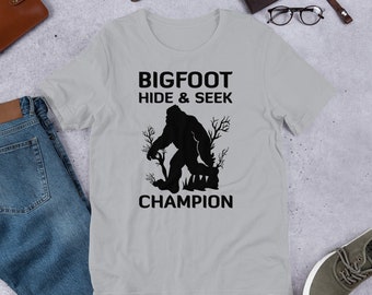 May include: A gray t-shirt with a black graphic of Bigfoot. The graphic says "Bigfoot Hide & Seek Champion".