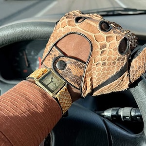 Op de afbeelding: Een gouden polshorloge met een vierkante wijzerplaat en een bruin leren bandje met een slangenhuidpatroon. Het horloge wordt gedragen aan de pols van een persoon, en de persoon draagt ​​een bruine leren handschoen met een slangenhuidpatroon.