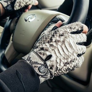 May include: A pair of fingerless driving gloves made of snakeskin with a black leather trim. The gloves have a button on the wrist with the letters "VI" on it.