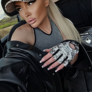 Puede incluir: Una mujer con una chaqueta de cuero negra, un top gris sin mangas y una gorra de béisbol con estampado de piel de serpiente. También lleva un par de guantes sin dedos con estampado de piel de serpiente.