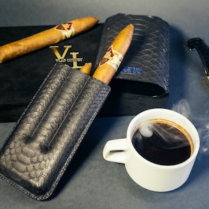 May include: Two brown cigars in a black leather cigar case with a snakeskin pattern. The case has the text "VLAD LUXURY" embossed on it. A white ceramic mug with black coffee and steam is in the background.