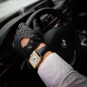 Puede incluir: Un reloj de oro con una esfera blanca y una correa de cuero negro. El reloj se lleva en la muñeca de una persona que lleva guantes de cuero negro y una camisa blanca. La persona está conduciendo un coche.