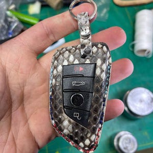 Op de afbeelding: Een zwarte en grijze autosleutelhouder met een slangenhuidpatroon en een zilveren sleutelring. De sleutelhouder heeft vier knoppen, waaronder een vergrendelingsknop, een ontgrendelingsknop, een kofferbakknop en een paniekknop. De sleutelhouder is omgeven door een witte, blauwe en rode stiksel.