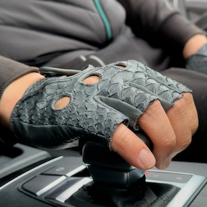 Puede incluir: Primer plano de un guante de conducir de cuero negro con un patrón texturizado. El guante está en la mano de una persona que está cambiando de marcha en un coche.