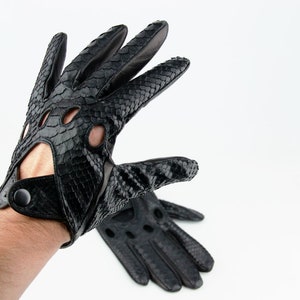 Puede incluir: Un par de guantes de conducir de cuero negro con un patrón de piel de serpiente. Los guantes tienen un cierre de botón en la muñeca y están hechos de cuero suave y flexible.