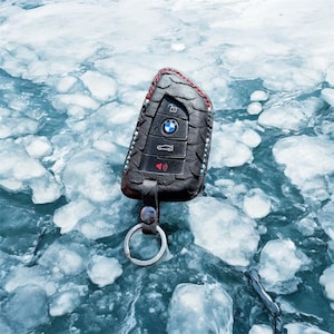 May include: A black leather key fob with a silver key ring. The key fob has a BMW logo and four buttons. The key fob is lying on a surface of ice.