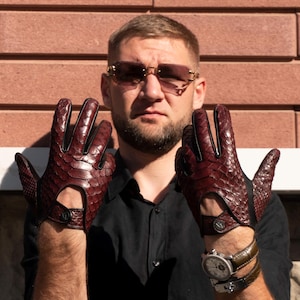 May include: A man wearing a black shirt and sunglasses is holding up a pair of brown leather driving gloves with a snakeskin pattern.