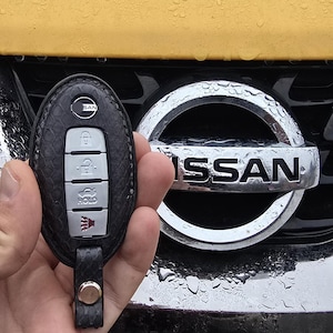May include: A black leather key fob with a silver Nissan logo and buttons. The key fob is held in a hand in front of a yellow car with a silver Nissan logo.