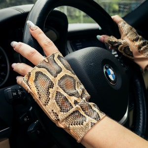 Puede incluir: Una persona lleva un par de guantes sin dedos de piel de serpiente marrón y beige mientras conduce un coche. Los guantes tienen un patrón texturizado y están hechos de cuero.
