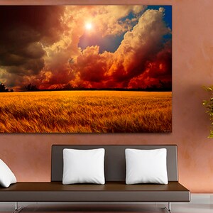 K&ouml;nnte beinhalten: Ein gro&szlig;er Leinwanddruck, der eine dramatische Landschaft darstellt. Das Kunstwerk zeigt ein goldenes Weizenfeld unter einem feurigen Himmel mit dunklen Wolken und einer hellen Sonne. Der Druck ist an einer Wand &uuml;ber einem braunen Sofa mit wei&szlig;en Kissen angebracht.