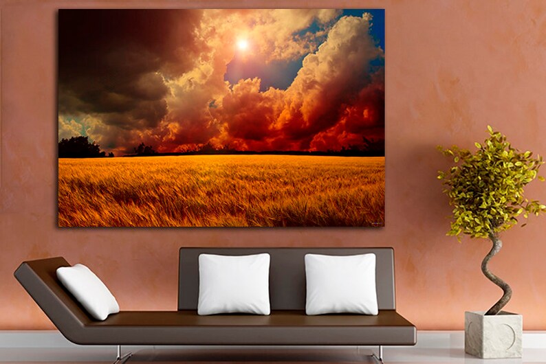 K&ouml;nnte beinhalten: Ein gro&szlig;er Leinwanddruck, der eine dramatische Landschaft darstellt. Das Kunstwerk zeigt ein goldenes Weizenfeld unter einem feurigen Himmel mit dunklen Wolken und einer hellen Sonne. Der Druck ist an einer Wand &uuml;ber einem braunen Sofa mit wei&szlig;en Kissen angebracht.