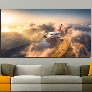Könnte beinhalten: Ein weißes Flugzeug fliegt über eine Bergkette mit Wolken und einem See darunter. Die Sonne geht im Hintergrund unter und taucht die Szene in ein goldenes Licht.
