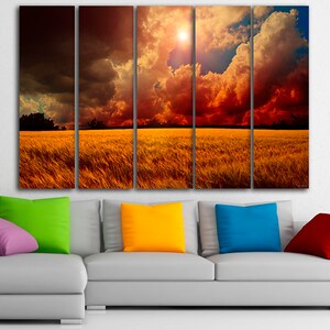 K&ouml;nnte beinhalten: Ein f&uuml;nfteiliger Leinwanddruck, der eine dramatische Landschaft zeigt. Das Kunstwerk zeigt ein goldenes Feld unter einem feurigen Himmel mit dunklen Wolken und einer hellen Sonne. Im Vordergrund befindet sich ein graues Sofa mit bunten Kissen in Lila, Gr&uuml;n, Gelb, Blau und Rot.