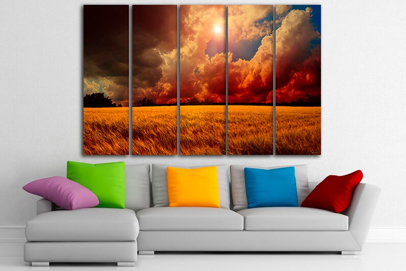 K&ouml;nnte beinhalten: Ein f&uuml;nfteiliger Leinwanddruck, der eine dramatische Landschaft zeigt. Das Kunstwerk zeigt ein goldenes Feld unter einem feurigen Himmel mit dunklen Wolken und einer hellen Sonne. Im Vordergrund befindet sich ein graues Sofa mit bunten Kissen in Lila, Gr&uuml;n, Gelb, Blau und Rot.