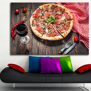 Puede incluir: Primer plano de una pizza de pepperoni con aceitunas verdes y albahaca sobre una mesa de madera con una copa de vino tinto, pimientos rojos y un mantel a cuadros rojo y blanco.