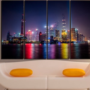 Könnte beinhalten: Eine nächtliche Skyline mit hohen Gebäuden und bunten Lichtern, die sich im Wasser spiegeln. Die Gebäude sind in verschiedenen Farben beleuchtet, darunter Rot, Gelb, Blau und Grün. Das Wasser ist ruhig und spiegelt die Lichter der Stadt wider.