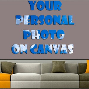 Op de afbeelding: Een grijs canvas met de tekst "YOUR PERSONAL PHOTO ON CANVAS" in blauwe letters met een achtergrond van lucht en wolken. Het canvas hangt aan een muur boven een grijze bank met oranje en groene kussens.