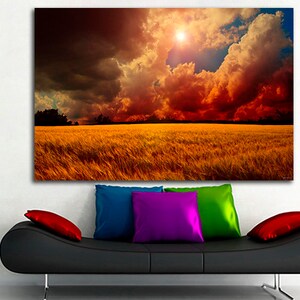 K&ouml;nnte beinhalten: Ein gro&szlig;er Leinwanddruck, der eine dramatische Landschaftsszene zeigt. Das Kunstwerk zeigt ein goldenes Feld unter einem feurigen Himmel mit dunklen Wolken und einer hellen Sonne. Der Druck h&auml;ngt &uuml;ber einem schwarzen Sofa mit bunten Kissen.