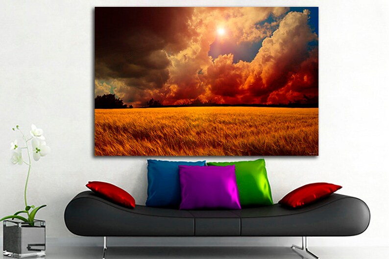 K&ouml;nnte beinhalten: Ein gro&szlig;er Leinwanddruck, der eine dramatische Landschaftsszene zeigt. Das Kunstwerk zeigt ein goldenes Feld unter einem feurigen Himmel mit dunklen Wolken und einer hellen Sonne. Der Druck h&auml;ngt &uuml;ber einem schwarzen Sofa mit bunten Kissen.