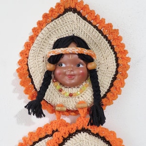 May include: Vintage wall hanging featuring a Native American doll face with black braids, a headband, and a beaded necklace. The doll is surrounded by a cream and orange crocheted design, with two matching coasters below. A decorative piece.