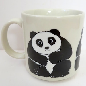 May include: Cream-colored ceramic mug featuring a black and white panda bear design. The panda is depicted in a seated position with a friendly expression. The mug has a curved handle and a cylindrical shape, suitable for hot beverages.