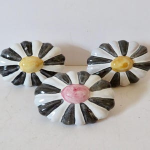 May include: Three ceramic daisy-shaped decorative objects. Each has a white base with black stripes, a yellow center, and a glossy finish. One daisy has a pink center. The objects are arranged on a white surface.