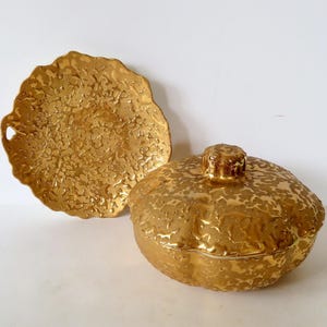 May include: A gold-coloured lidded serving dish with a textured, organic surface pattern alongside a matching round plate featuring a scalloped edge and a small side handle. Both items exhibit a metallic finish with a consistent, raised, mottled texture.
