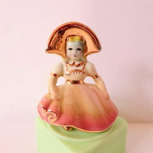 May include: A porcelain figurine of a girl wearing a pink and yellow dress and a gold hat. The figurine is standing on a green base.