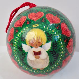 May include: A round Christmas ornament with a red cord for hanging. The ornament features a green background with red hearts and a central illustration of an angel with a halo, wings, and a book. The angel has yellow hair and is wearing a white robe.