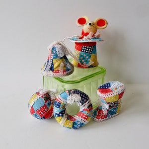 May include: A set of five patchwork fabric ornaments, including a bell, a ball, a ring, and two other shapes. The ornaments are decorated with red, blue, white, and yellow fabric squares. A small mouse figurine is sitting on top of a patchwork fabric hat.