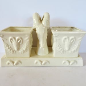 May include: A cream-colored ceramic double planter featuring two square containers connected by a central ram head sculpture. Each square container displays a relief of a ram head and foliage. The base is rectangular with small decorative arches.
