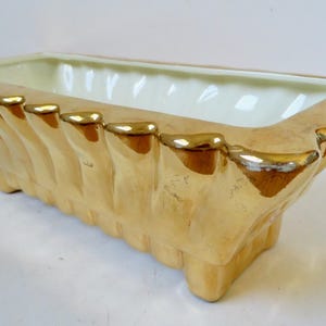 May include: A rectangular, ceramic planter with a cream interior and a light tan exterior. The planter has a scalloped design with gold trim along the top edge. The planter is approximately 12 inches long, 5 inches wide, and 4 inches tall.